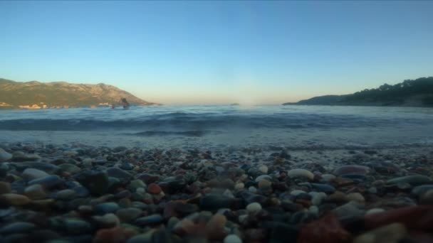 Spiaggia di ghiaia e rocciosa della città di Becici vicino Budva sul tramonto. Resort. Le onde del mare coprono la telecamera e va sott'acqua. I bambini nuotano nel mare. Viaggio turistico e viaggi. Umore romantico. Vacanza. — Video Stock