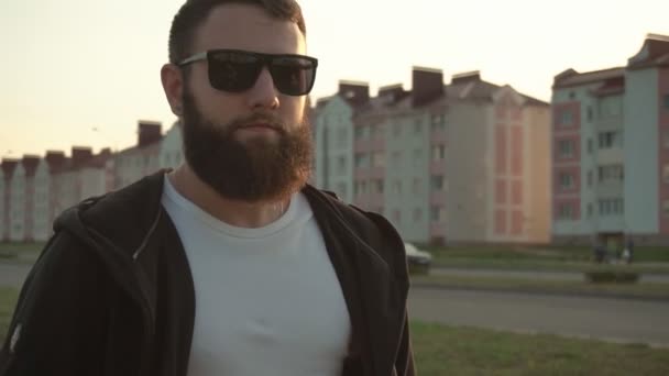 A handsome young caucasian man with a bushy beard in black glasses walks in the weather, background. Sunny sunset, slow motion, model — Stock Video