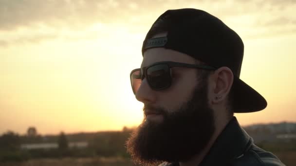 Ein gutaussehender brutaler Mann mit buschigem Bart und schwarzer Brille steht vor einem Sonnenuntergang. Hintergrund, Zeitlupe, modern — Stockvideo