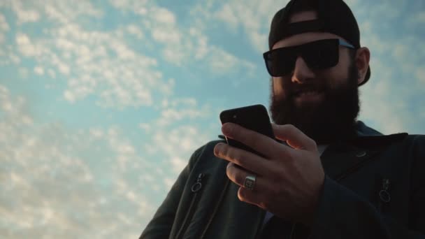 Stylischer brutaler Mann mit schwarzer Brille und Bart schaut auf das Telefon und lacht vor blauem Himmel, Zeitlupe, modisch — Stockvideo