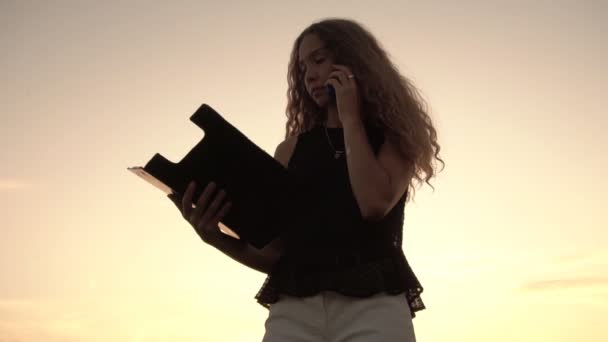 Biała kobieta biznesu rozmawia przez telefon na tle słonecznego zachodu słońca i trzyma tabliczkę z papierami w ręku. Zwolniony ruch — Wideo stockowe