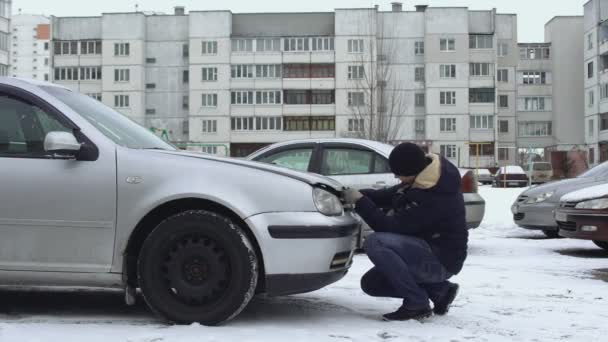 Человек пытается и не может открыть капот своей машины зимой на парковке двора. Капюшон замерз из-за холодной погоды. Срыв. Неисправность или проблема с автоматикой. Хочется зарядить батарею — стоковое видео