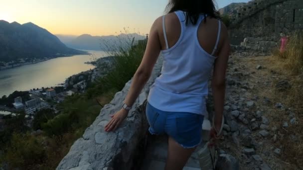Όμορφη κοπέλα τουρίστρια βόλτες το βράδυ μέσα από τα βουνά του Μαυροβουνίου στην πόλη Kotor. Κορίτσι θαυμάζει το ηλιοβασίλεμα με θέα τα βουνά και τη λίμνη, αργή κίνηση, διακοπές — Αρχείο Βίντεο