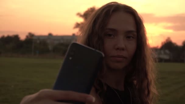 Mooi meisje met krullend haar maakt een selfie op de telefoon tegen de achtergrond van een zonnige zonsondergang, slow motion — Stockvideo