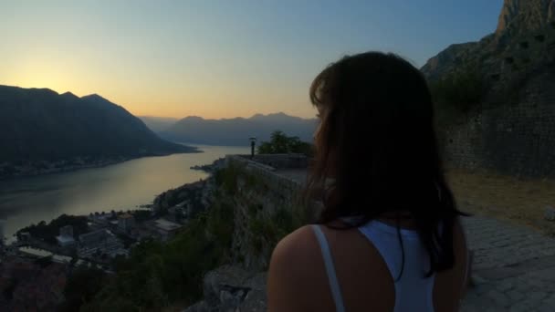 Ein kaukasisches Mädchen bewundert den Sonnenuntergang von einer Höhe über den Bergen und dem See. große touristische Spot in der Stadt kotor. Abendsonnenuntergang mit Bergblick, Stadtbeleuchtung, Zeitlupe, Reise — Stockvideo