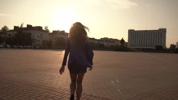 Una hermosa chica con el pelo rizado largo corre alrededor de la ciudad y se da la vuelta, mira a la cámara contra la puesta de sol, lento-mo — Vídeo de stock