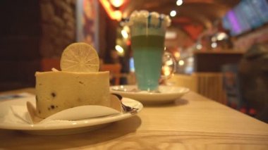 Masada nefis peynirli kek ve bardakta kokteyl. Kafede rahat bir atmosfer ve ziyaretçiler, fotokopi alanı, tatlı