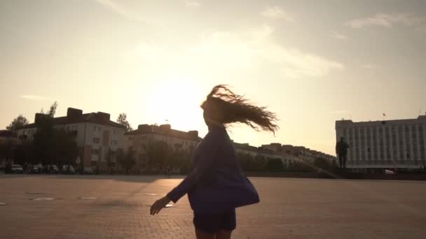 Ein schönes Mädchen mit langen lockigen Haaren läuft durch die Stadt und dreht sich um, blickt in die Kamera gegen den Sonnenuntergang, — Stockvideo