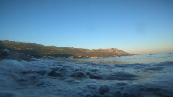 Ondas no mar lavar a costa de pedras e seixos contra o pano de fundo de montanhas de céu azul e montanhoso. O conceito de descanso e relaxamento, espaço de cópia, câmera lenta — Vídeo de Stock