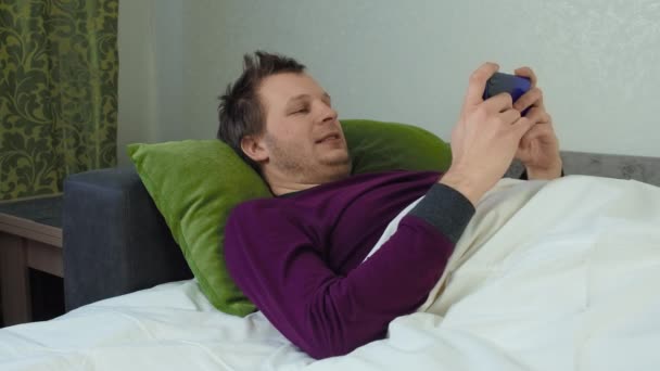 El hombre cubierto con una manta está acostado en la cama y jugando un juego móvil. Concepto de insomnio, insomnio. Un caucásico sin afeitar. Bloqueo a mitad de disparo — Vídeo de stock