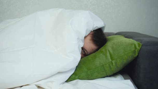 Ο άνθρωπος είναι ξαπλωμένος στο κρεβάτι και κοιτάζει έξω κάτω από την κουβέρτα και κρύβεται πίσω. Το κεφάλι του άντρα είναι ξαπλωμένο στο μαξιλάρι. Περίεργα καυκάσια στραβάδια και γέλια. Περίεργη κατάσταση. Κλείδωμα μέσης βολής — Αρχείο Βίντεο