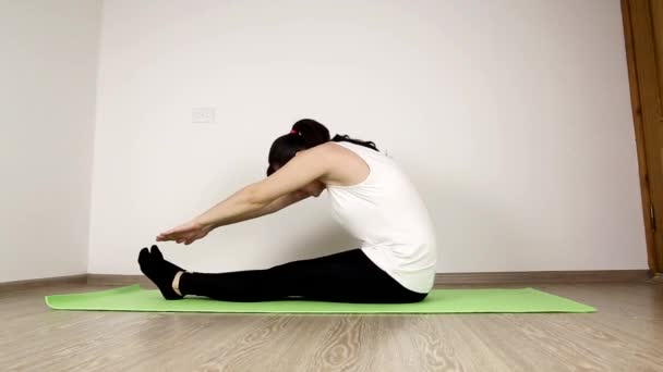 Joven hermosa morena caucásica chica en una camiseta blanca hace gimnasia en casa. Realiza un ejercicio de estiramiento estirado por las manos hasta los dedos de los pies, flexibilidad — Vídeos de Stock