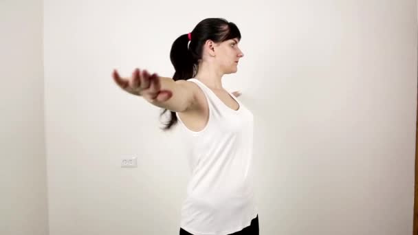 Νεαρή όμορφη μελαχρινή καυκάσιος κορίτσι σε λευκό μπλουζάκι κάνει γυμναστική στο σπίτι. Εκτελεί μια άσκηση ταλάντευσης βραχίονα για το στήθος, υγιή — Αρχείο Βίντεο