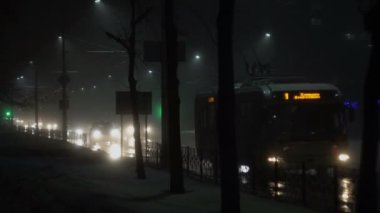 Caddede arabalar, araçlar, toplu taşıma araçları sürüyor. Kışın sisli bir gece şehri. Islak asfalta yansıyor. Farlardan gelen ışık. Yol trafiği ve kötü hava için görüş mesafesi düşük.