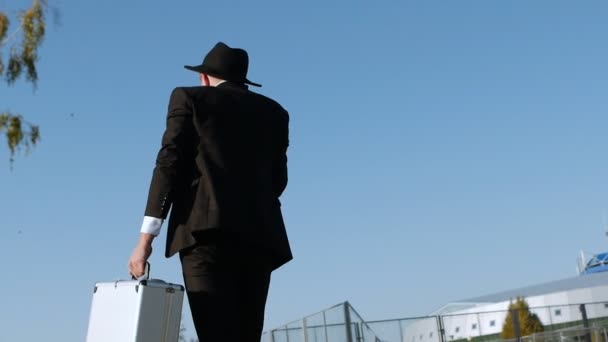 El hombre de traje negro y sombrero con estuche plateado va a la reunión para transferir dinero. Concepto de corrupción y soborno. Vista trasera, detrás. Hombre de negocios parece judío o mafia italiana está hablando en el teléfono inteligente . — Vídeos de Stock