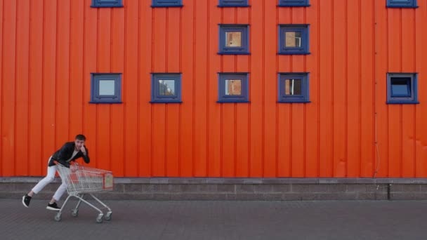 幸せな顧客の男は外のモール店の中心の壁に沿って空のショッピングカートに乗り、親指を上げます。割引や販売の概念.面白いと遊園地の瞬間 — ストック動画