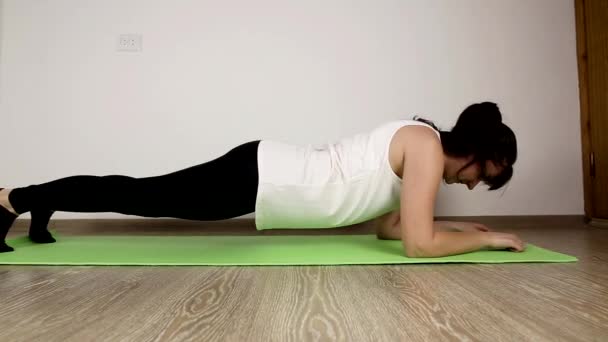 Menina branca morena bonita nova em um top de tanque branco faz ginástica em casa. Executa uma prancha de exercício, fundo — Vídeo de Stock