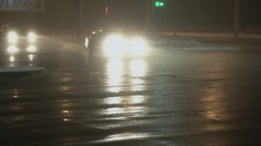 Arabalar, araçlar trafik ışıklarıyla kavşaktan geçiyor. Kışın sisli bir gece şehri. Ağır çekim. Islak asfalta yansıyor. Farlardan gelen ışık. Yol trafiği ve kötü hava için görüş mesafesi düşük.