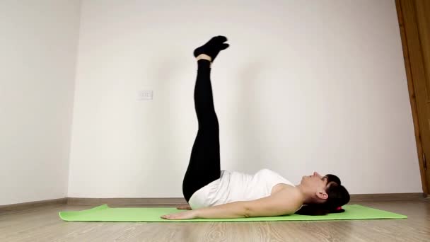 Menina branca morena bonita nova em um top de tanque branco faz ginástica em casa. Realiza exercício de levantamento de perna para fortalecer os abdominais — Vídeo de Stock