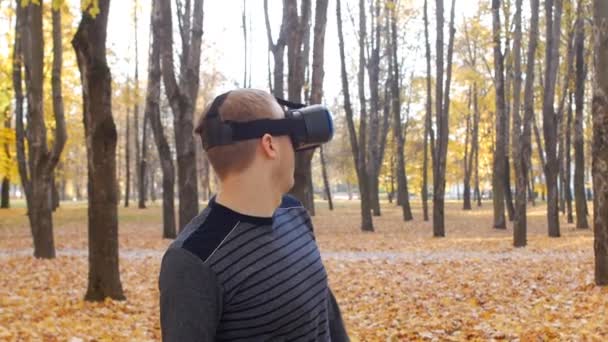 Um jovem caucasiano em óculos de realidade virtual examina o espaço ao redor. Bela natureza, parque de outono, câmera lenta, jogador — Vídeo de Stock