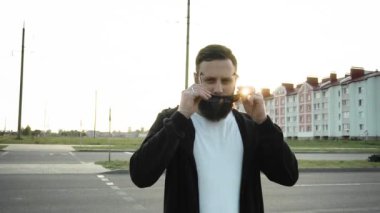 Sakallı, modaya uygun bir adam siyah gözlük takıyor. Vahşi stil sahibi adam gün batımında ağır çekimde duruyor. Küpeli, yüzüklü ve parmaklarında mühür olan modaya uygun biri. Şık bir moda.