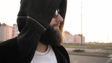 Vahşi stil sahibi adam gün batımında gülümsüyor ve ağır çekimde duruyor. Siyah gözlüklü ve kapüşonlu sakallı, modaya uygun bir adam. Modayı takip eden biri. Moda insanları kavramı. Yaşam tarzı