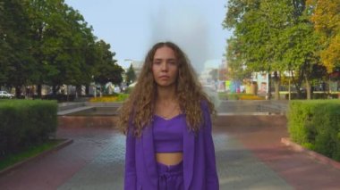 Makyajsız, beyaz, kıvırcık saçlı, şirin bir kızın portresi. Modaya uygun bir kadın ağır çekimde hareketsiz duruyor. Moda düşkünü bayan parkta. Vogue dişisi kameraya bakar