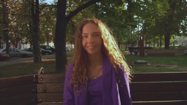 Vogue curly long haired girl in a suit without makeup sits on bench in city park under sun rays, looks into a camera. Natural beauty without cosmetics. Fashionable stylish woman. Trendy fancy lady — Stock Video
