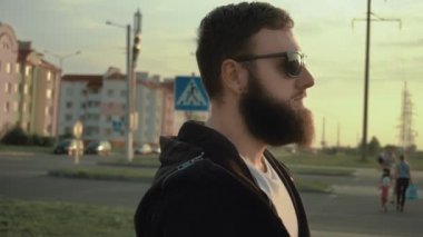 Siyah gözlüklü şık sakallı adam gün batımında şehir caddesinde ağır çekimde yürüyor. Küpeli acımasız bir adam. Siyah cüppeli modaya uygun biri. Konut alanı. Sabit çekim