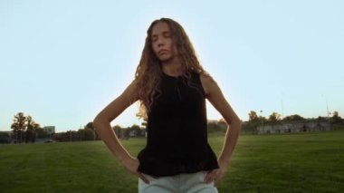 Makyajsız, beyaz, kıvırcık saçlı bir kızın portresi. Gün batımı. Vogue modeli dışarıda poz veriyor. Modaya uygun bir kadın kameraya bakar, güneş ışınları altında ağır çekimde durur..