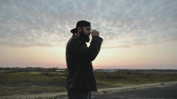 El tipo brutal barbudo se pone gafas negras al atardecer en cámara lenta. Hombre elegante de moda en gorra y chaqueta de cuero se detiene. Persona elegante de moda con pendiente. Chic modish Estilo de vida. Biker. — Vídeo de stock