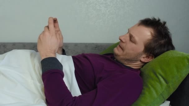Ένας νεαρός αξύριστος καυκάσιος παίζει στο τηλέφωνο ξαπλωμένος στο κρεβάτι. Ένας τύπος gamer παίζει ένα παιχνίδι σε ένα smartphone. — Αρχείο Βίντεο