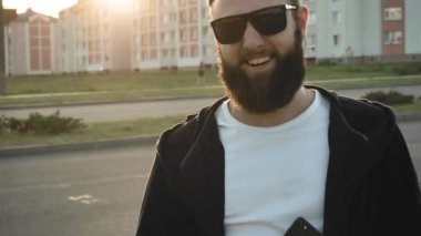 Siyah gözlüklü mutlu adamın sakallı portresi. Vahşi stil sahibi adam gün batımında ağır çekimde gülümser. İnsani samimi gerçek duygular. Kulağında küpe olan beyaz adam şehirde hareketsiz duruyor.