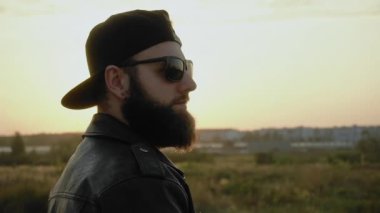 Siyah gözlüklü, şapkalı, deri ceketli, son moda adam gün batımında. Modaya uygun bir adam ağır çekimde hareketsiz duruyor. Küpeli süslü bir insan. Şık, modaya uygun. Yaşam tarzı. Motorcu