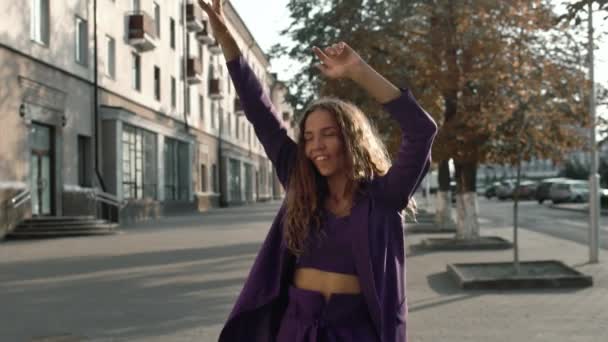 Feliz encaracolado longo cabelo sorridente menina dançando ao ar livre na rua da cidade. A mulher elegante na moda no terno move-se no movimento lento. Conceito positivo. Vogue fêmea olha para uma câmera. Senhora de negócios na moda — Vídeo de Stock