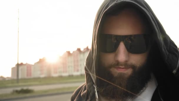 El barbudo con gafas oscuras y capucha mira a una cámara al atardecer. Brutal hombre con estilo se detiene en cámara lenta. Persona de moda de moda. Estilo de vida, gente caucásica. Copiar espacio. Steadicam tiro — Vídeos de Stock