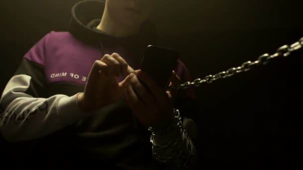 Een man houdt een smartphone in zijn hand waaraan een ketting is bevestigd. Smartphone en internet verslaving concept onder tieners, achtergrond, slow mo — Stockvideo
