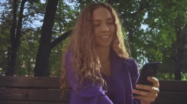 Gülümseyen kıvırcık saçlı, makyajsız kız akıllı telefona bakıyor ve haberleri izliyor. İletişim, sohbet, sosyal ağ, mesajlaşma. Kadın vlogger sörfü ve Wi-fi ya da 4g internet üzerinden gezinme