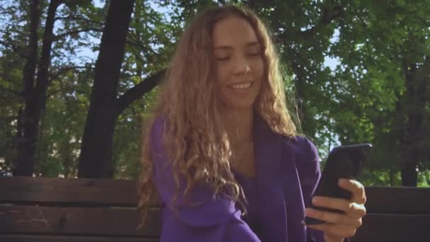 Smiling Curly long haired girl without makeup looks at smartphone and watching news feed. Communication, chatting, social networking, messager. Female vlogger surf and browse via wi-fi or 4g internet — Stock Video