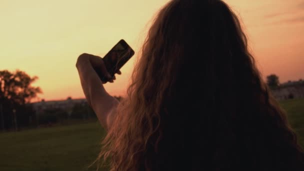 Fille blogueuse aux cheveux longs bouclés prend des photos selfie au téléphone au coucher du soleil pour ses abonnés ou abonnés dans le service de réseautage social. Vue arrière à l'arrière. Vogue femelle regarde dans la caméra smartphones .