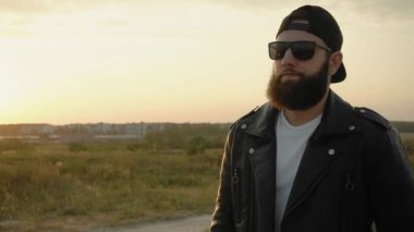 Siyah gözlüklü, şapkalı, deri ceketli, vahşi sakallı adam gün batımında yavaş çekimde dışarı çıkıyor. Modaya uygun bir adam ağır çekimde gider. Küpeli modaya uygun biri. Vogue yaşam tarzı. Motorcu