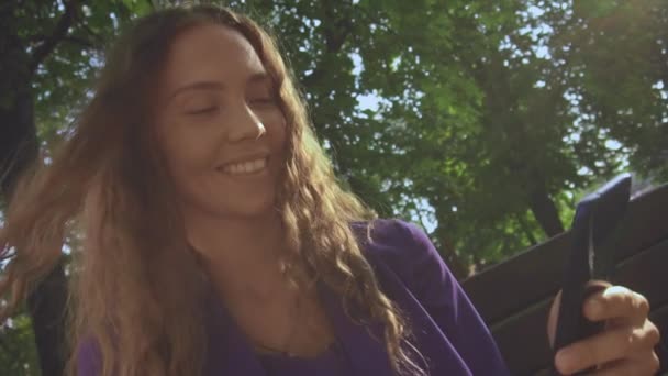 Sonriendo rizado chica de pelo largo. El pelo vuela con viento bajo los rayos del sol. Mujer sin maquillaje mira el teléfono inteligente y ver noticias de alimentación. Comunicación, redes sociales, mensajero. Vlogger mujer — Vídeos de Stock