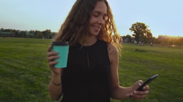 Souriante fille bouclée aux cheveux longs avec une tasse de café et un smartphone au coucher du soleil dans le parc en regardant les actualités. Femme sans maquillage passe commande dans la boutique en ligne et effectue le paiement d'achat. Achats à distance — Video