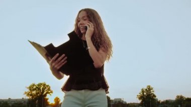 Vogue kadın modelleri dışarıda kendi işlerine kendileri karar verir. Kıvırcık saçlı, elinde klasörle günbatımında telefonda konuşan iş kadını. Beyaz kadın ağır çekimde hareketsiz duruyor.