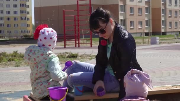 En vacker ung mor leker i sandlådan med sin lilla dotter. Leka på lekplatsen en solig dag, utomhus — Stockvideo