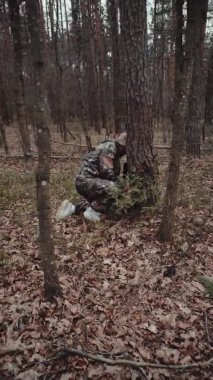 Dikey çekim. Kamuflaj kıyafeti giymiş avcı orman bekçisi bir ağacın arkasında bir şey saklıyor. Garip muhabir. Panama şapkalı ve bıyıklı blog yazarı