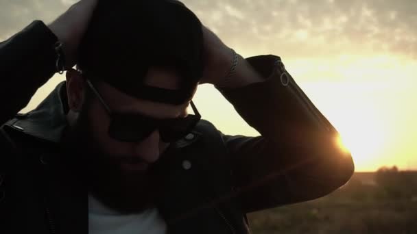 Barbudo motociclista elegante se pone la gorra negra al atardecer en cámara lenta. Hombre elegante de moda en gafas y chaqueta de cuero. Brutal persona de moda con pendiente. Chic modish Estilo de vida. Biker. . — Vídeo de stock