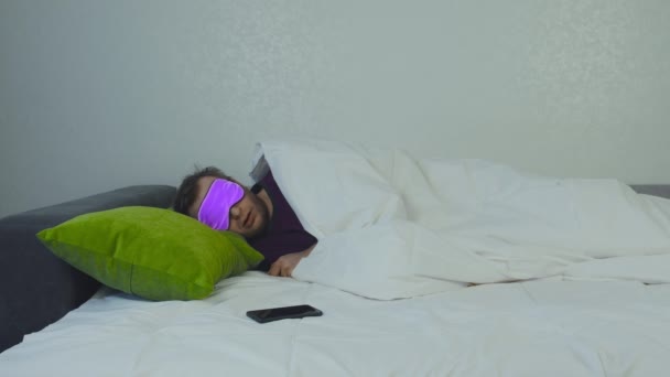 Красивый белый мужчина в маске для сна спит на кровати, рядом лежит смартфон. Здоровая концепция сна человека, фон — стоковое видео