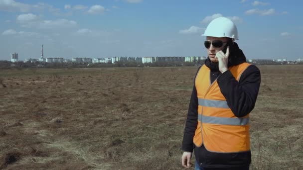 Engenheiro construtor falando por telefone sobre novas obras de construção em campo. Chefe chefe em capacete branco, colete laranja, em óculos pretos gestos acenando com as mãos. Mercado imobiliário . — Vídeo de Stock