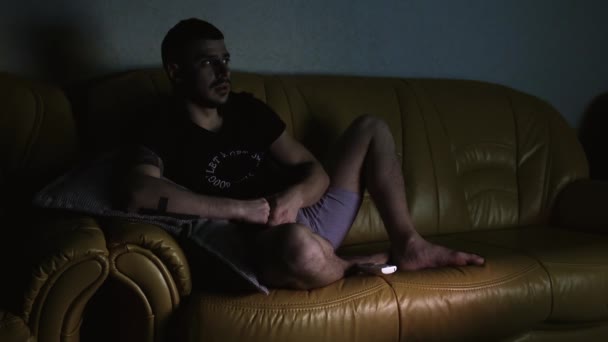 El tipo se sienta en casa solo en el sofá y ve películas de terror en la televisión por la noche en el apartamento. Pasar tiempo en cuarentena de auto-aislamiento. Joven hombre caucásico de 30 años con bigote vistiendo camisa y pantalones cortos — Vídeo de stock